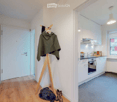 A narrow hallway leads to a modern kitchen area with white cabinets, a built-in oven, and a sink. A coat rack with a jacket and shoes is visible in the hallway.