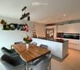 Modern kitchen with white cabinets, stainless steel appliances, a wooden dining table with six chairs, and a flower vase on the table. The kitchen features a pendant light fixture and a decorative wall with flags.