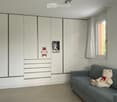 A furnished room with a grey sofa, a large white wardrobe, and a teddy bear toy on the sofa. The room has a window with white curtains and a small photo on the wardrobe.