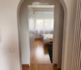 A hallway with wooden flooring and white walls, leading to a bedroom with a bed, rug, and window with curtains.