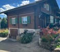 Two-story wooden house with green shutters, a garage, and a well-maintained garden with stone pathways and decorative plants.