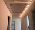 Empty hallway with light wooden flooring, white walls, and recessed lighting. Doors lead to adjacent rooms, one with a window and blinds visible.
