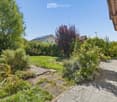 Well-maintained garden with a variety of plants, shrubs, and trees, featuring a paved pathway leading to a wooden bench near the house.