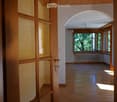 Empty hallway with wooden floors, white walls, large windows, and a radiator. The hallway features an arched ceiling and a wooden door with glass panels on the left side.