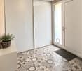 A modern hallway with a decorative hexagonal tile floor, white walls, recessed lighting, and a potted plant on a console table. Features a sliding glass door leading outside.