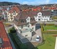 Aerial view of a residential neighborhood with multiple houses, red-tiled roofs, and a mix of architectural styles. A white car is parked in a driveway, and there is a small garden area with a swimming pool and potted plants.