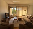 Bright living room with wooden flooring, large window with curtains, sofa, coffee table, ottoman, armchairs, TV stand, and books.