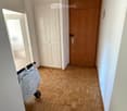 A narrow hallway with wooden herringbone flooring, white walls, and a hanging woven basket light fixture. Features a wooden door on the right and a white double door on the left, with a stroller visible in the foreground.