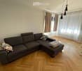 Living room with a grey sectional sofa, patterned pillows, wooden floor, large windows with curtains, radiator, and modern pendant lights.