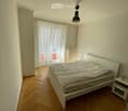 Minimalist bedroom with a white bed, nightstand, and wooden flooring. Large window with white curtains allows natural light to enter.