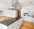 Modern kitchen with white cabinets, granite countertops, induction cooktop, built-in oven, and range hood. Wooden flooring and large window with view of greenery.