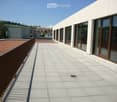 Modern rooftop terrace with light grey tiled flooring, metal railing, and large windows overlooking a residential area.