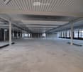 Empty industrial-style room with concrete floor, exposed white beams, and large windows allowing natural light.