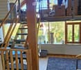 The image shows a modern, open-concept living space with wooden beams and large windows. Visible objects include a staircase, a cat tree, a laptop, a fan, a radiator, a table, a chair, and a toy car. The room features a mix of wooden and tiled flooring, w