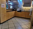A well-lit kitchen with wooden cabinets, a tiled floor, and various appliances including a microwave, coffee maker, and oven. There is a sink with a faucet and a towel hanging nearby.
