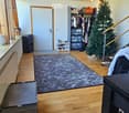 A spacious, well-lit room with wooden flooring, a large grey rug, a black speaker, a wardrobe, a shoe rack, a Christmas tree, and a small table with a chair.
