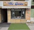Facade of a gelateria named "Me Gusta" with a striped awning, glass display window, and menu board listing soft drinks and coffee options.