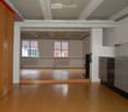 An unfurnished room with wooden flooring, white walls, and exposed wooden beams on the ceiling. A kitchen area is visible with cabinets and a sink, but no furniture or appliances are present.