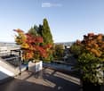 Modern balcony with tiled flooring, metal railing, and potted plants, overlooking a cityscape with trees and parked cars.
