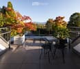 Balcony with a round table and four black metal chairs, surrounded by trees and a clear view of the street.