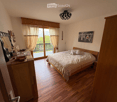 Cozy bedroom with wooden flooring, large bed with checkered bedding, nightstand, dresser with mirror, artwork on wall, and large window with curtains offering a scenic view.