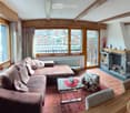 Cozy living room with wooden beams, large windows offering a scenic view, a sectional sofa, coffee table, ottoman, dining table with chairs, television, fireplace, and decorative items.