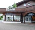 A two-story wooden building with a balcony and large windows, featuring a storefront with glass display windows and signage reading "Fleur de Pierre" and "Edelkreationen".
