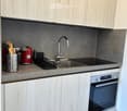 Modern kitchen with light wood cabinets, stainless steel appliances, and a grey countertop. Features include a sink with a modern faucet, an oven, and various kitchen utensils on the counter.
