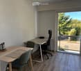 A modern, well-lit room with a desk, office chair, small table, and a laptop, featuring large windows with a view of greenery.