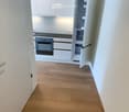 Empty hallway with wooden flooring leading to a modern kitchen area featuring white cabinets, a built-in oven, and a refrigerator. The kitchen is clean and unfurnished.