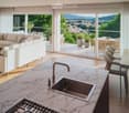 Modern, open-concept living and dining area with a view of the city and mountains. Features a large sofa, coffee table, armchair, kitchen island with sink and faucet, dining table with chairs, potted plants, and a television on a cabinet.