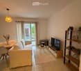 Modern living room with a white sofa, coffee table, TV stand, and a small dining area. Features a balcony with outdoor furniture visible through large glass doors.