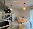 A compact, well-lit studio apartment with a combined living and sleeping area. Features include a sofa, small dining table with chairs, a bed, a refrigerator, washing machine, microwave, and a kitchen island with an oven. The space is decorated with a vas