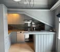 Modern kitchen with light grey cabinetry, a central island, and stainless steel appliances including a stove and dishwasher. The space features a sleek, minimalist design with a hanging pendant light fixture above the island.