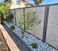 Modern garden area with a small tree, shrubs, white pebbles, and a low concrete wall, bordered by a grey privacy fence.
