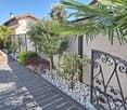 A well-maintained garden area with a paved walkway, decorative white pebbles, small trees, shrubs, and a wrought-iron railing leading to a house entrance.