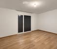 Empty room with wooden flooring, white walls, and large sliding glass doors with black blinds.