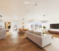 Open-concept living and dining area with hardwood floors, large windows offering a city view, a grey sofa, coffee table, dining table with chairs, kitchen island, pendant lights, and a television.