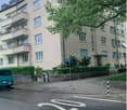Multi-story residential building with multiple windows and balconies, surrounded by greenery and a parked van.