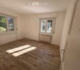 Empty room with wooden flooring, white walls, and large windows allowing natural light to enter. There is a radiator under one window and a door visible on the right side.