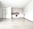 Empty room with tiled floor, white walls, and a closed door. Kitchen area visible with cabinets and appliances.