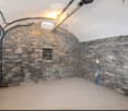 Empty basement room with exposed stone walls, concrete floor, and visible pipes and electrical wiring. No furniture or furnishings present.