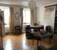 A well-lit room with wooden flooring, large windows with curtains, a sofa, a table, a bookshelf, a mirror, a lamp, and a radiator.