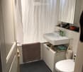 Modern bathroom with dark tiled floor, white sink, toilet, mirror, and a towel hanging on the wall. A shower curtain is visible, and a small basket and stool are placed near the door.