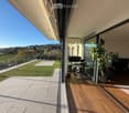 Spacious balcony with wooden flooring, large glass sliding doors, modern furniture including a desk and office chair, potted plants, and a small artificial grass area with lounge chairs.