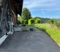 Driveway leading to a wooden house with a covered entrance, surrounded by greenery and a few potted plants.