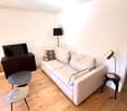 Modern living room with light wooden flooring, white walls, and a white sofa with patterned cushions. Includes a coffee table, two side tables, a floor lamp, and a television on a stand.
