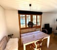Brightly lit room with wooden floor, large window, wooden bench with fur throw, dining table with chairs, small black stool, and TV on stand.