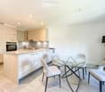 Modern kitchen with light wood cabinetry, a central island, and a glass dining table with four chairs. The space includes an oven, sink, and a floor lamp near the dining area.