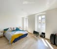 Bright bedroom with wooden flooring, large windows, a neatly made bed with colorful bedding, a nightstand, a laptop, a screen, and two suitcases near the window.